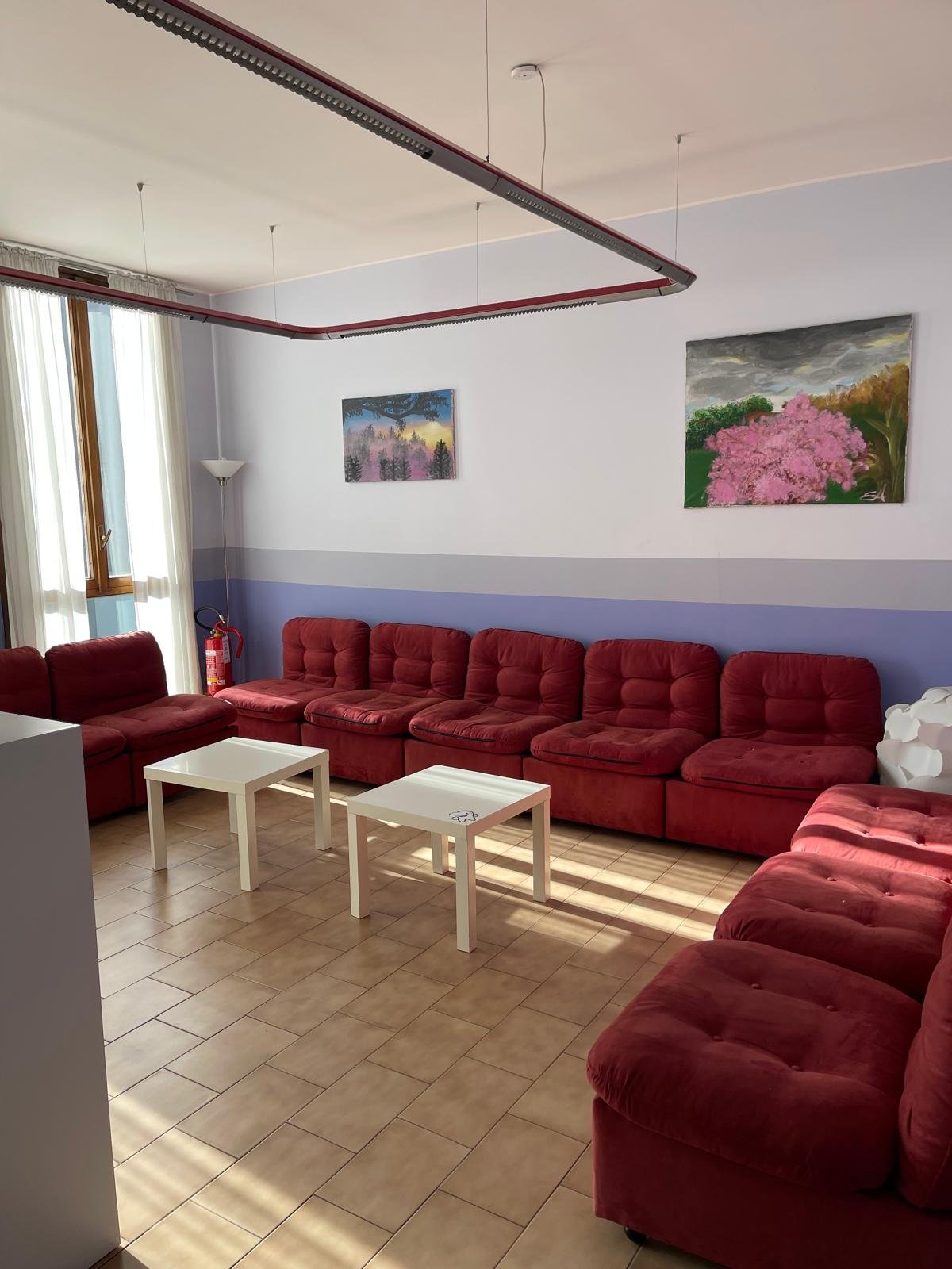 Sala di soggiorno con divani rossi, due tavolini bianchi, pareti bianche con una fascia azzurra, quadri floreali alle pareti, finestra con tende chiare, lampada da terra.