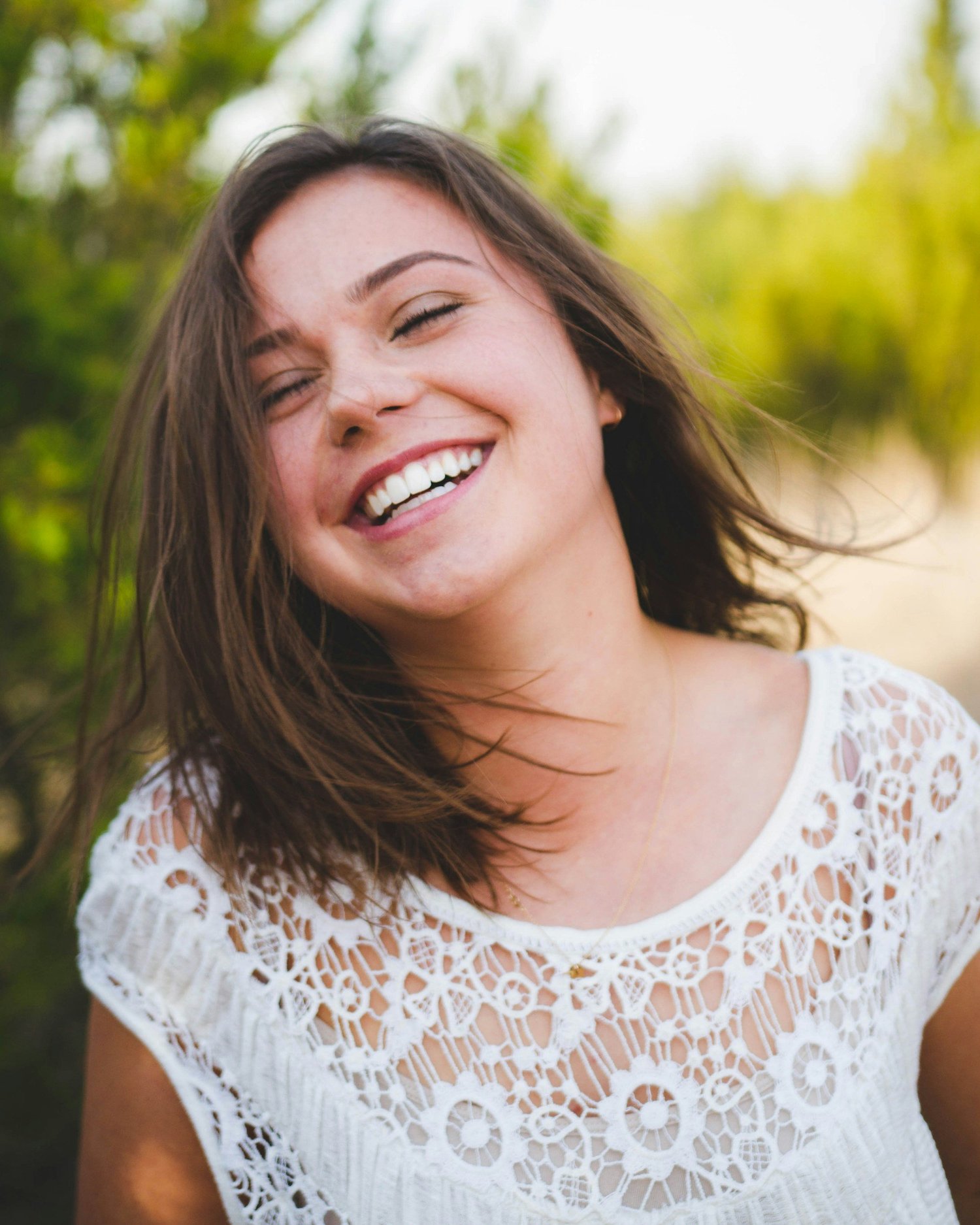 Ragazza sorridente con capelli mossi, abbraccia il cielo, sfondo di natura verdeggiande.